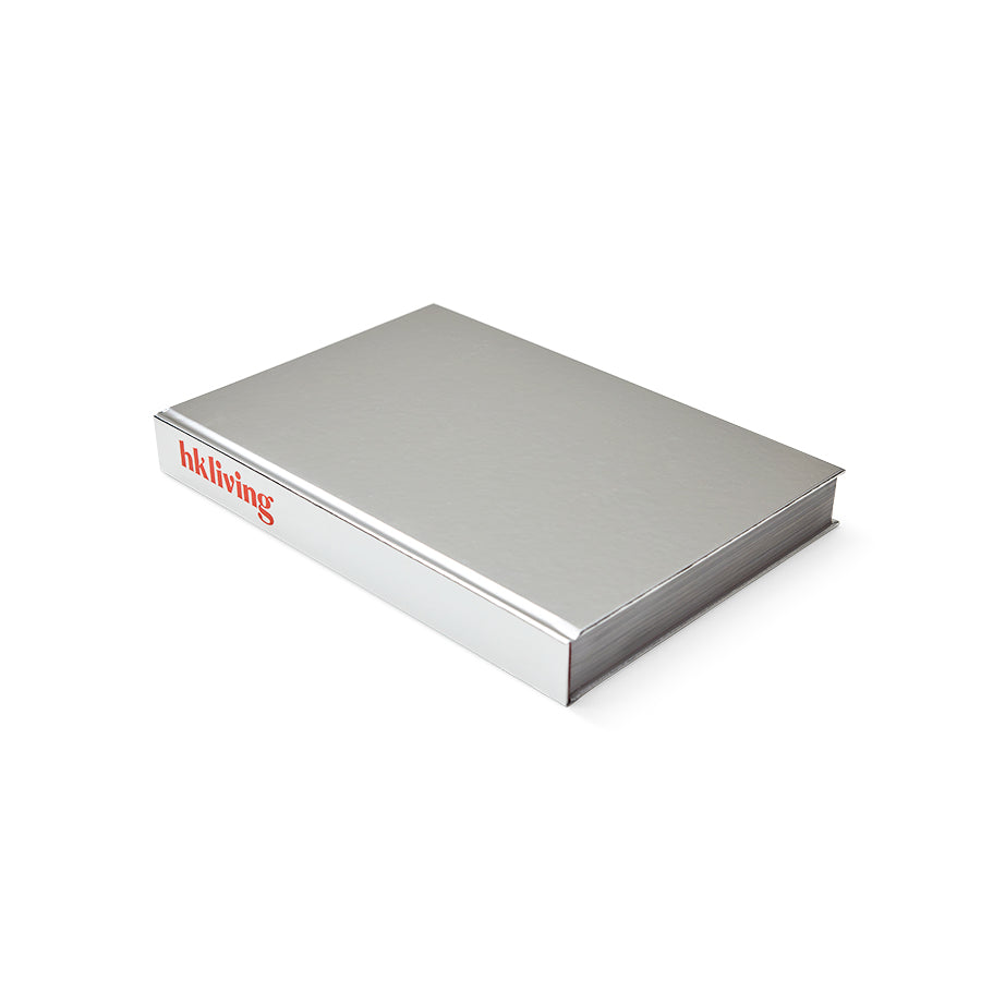 HKLIVING Limited Edition Lookbook 2026-2027, closed hardback book with mirrored silver foil cover and red HKLIVING logo mark, angled view showing the spine