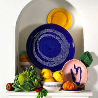 a large serving plate centres this image surrounded by other plates and bowls from the ottolenghi from serax range showing off yellow, pink, aure, and deep blue along with vegetables and fruits common in ottolenghi's recipes