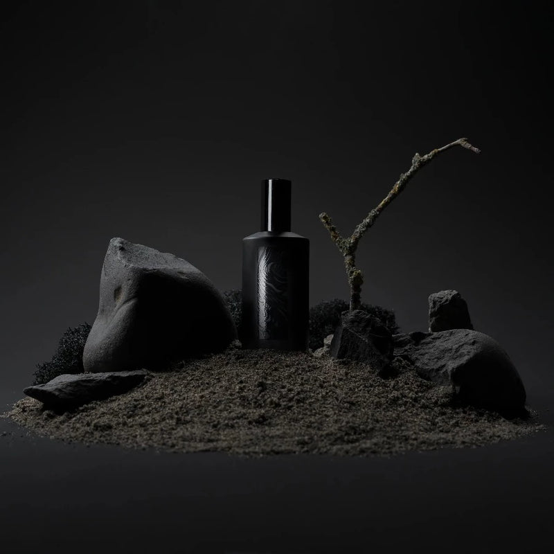 a black bottle of flotholt perfume stood on a sandy hill with black rocks around it