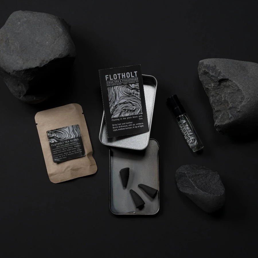 the fischersund flotholt scent survival kit showing the open tin which contains a small spray of the eau de parfum, incense cones, and tea with nroken black rocks around