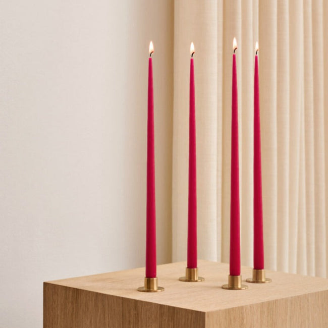 4 tall red taper candes in gold holders standing alight on a square, wooden table