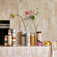 different hkliving mugs and vases stacked and stood on a tiled shelf in a kitchen with individual stems of flowers in each of the vases