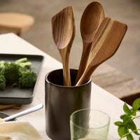 Aida wooden utensils in a black container on a kitchen counter with vegetables and a glass.