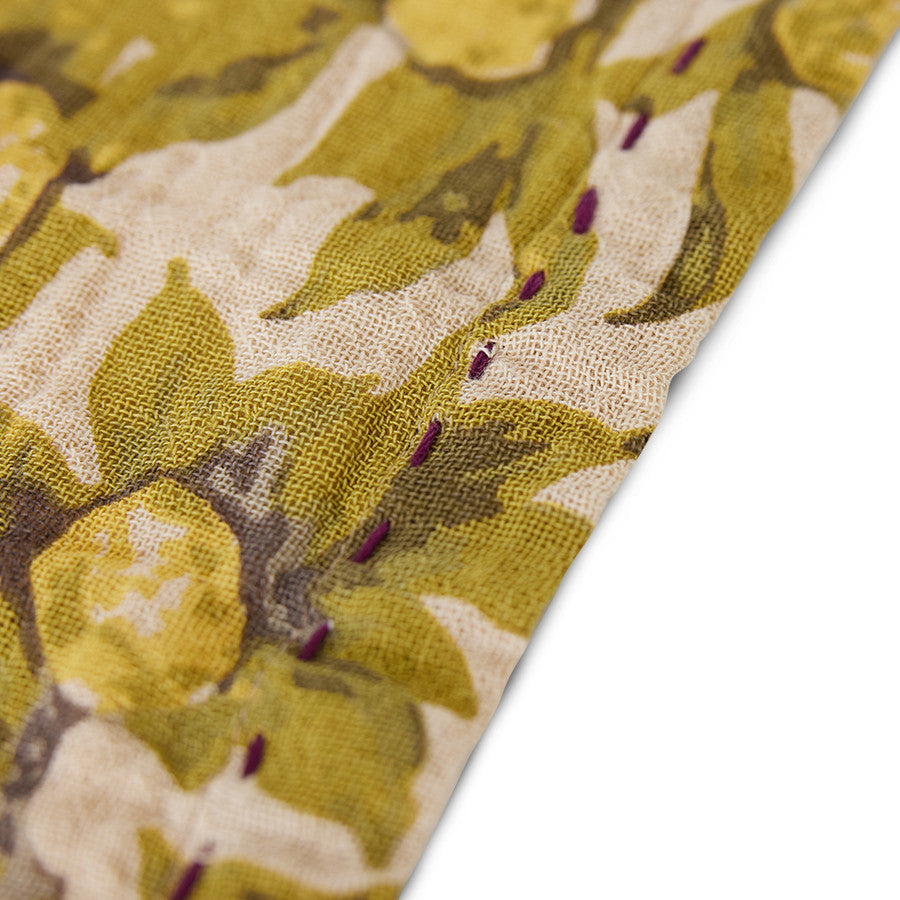 close up of the corner of a cloth napkin from hkliving with a fruiting branch style in yellow, green, and beige, with a purple stitching around the outeredge