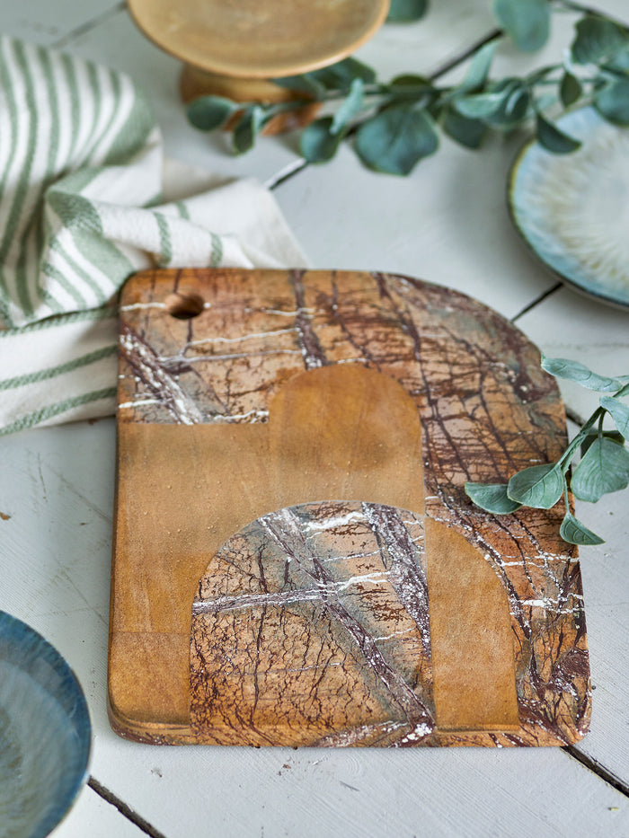 Brown marble cutting board styled on a rustic kitchen table with greenery, dishes, and natural textures.