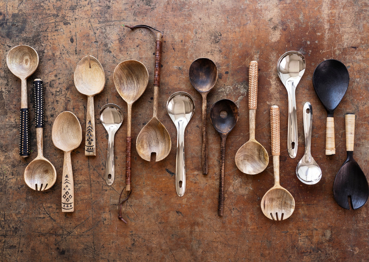 assorted handcrafted wooden and metal serving spoons on a rustic tabletop including dark mango wood servers
