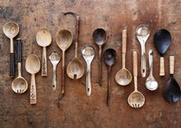 assorted handcrafted wooden and metal serving spoons on a rustic tabletop including dark mango wood servers
