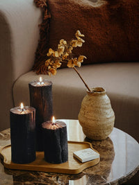 Three lit black Coal Figure Candles in different heights on a tray beside a woven vase and dried branches