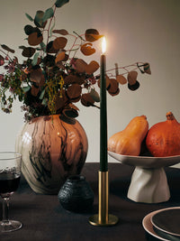 Brass Maro candle holder with a lit dark green taper beside a marbled vase and fruit