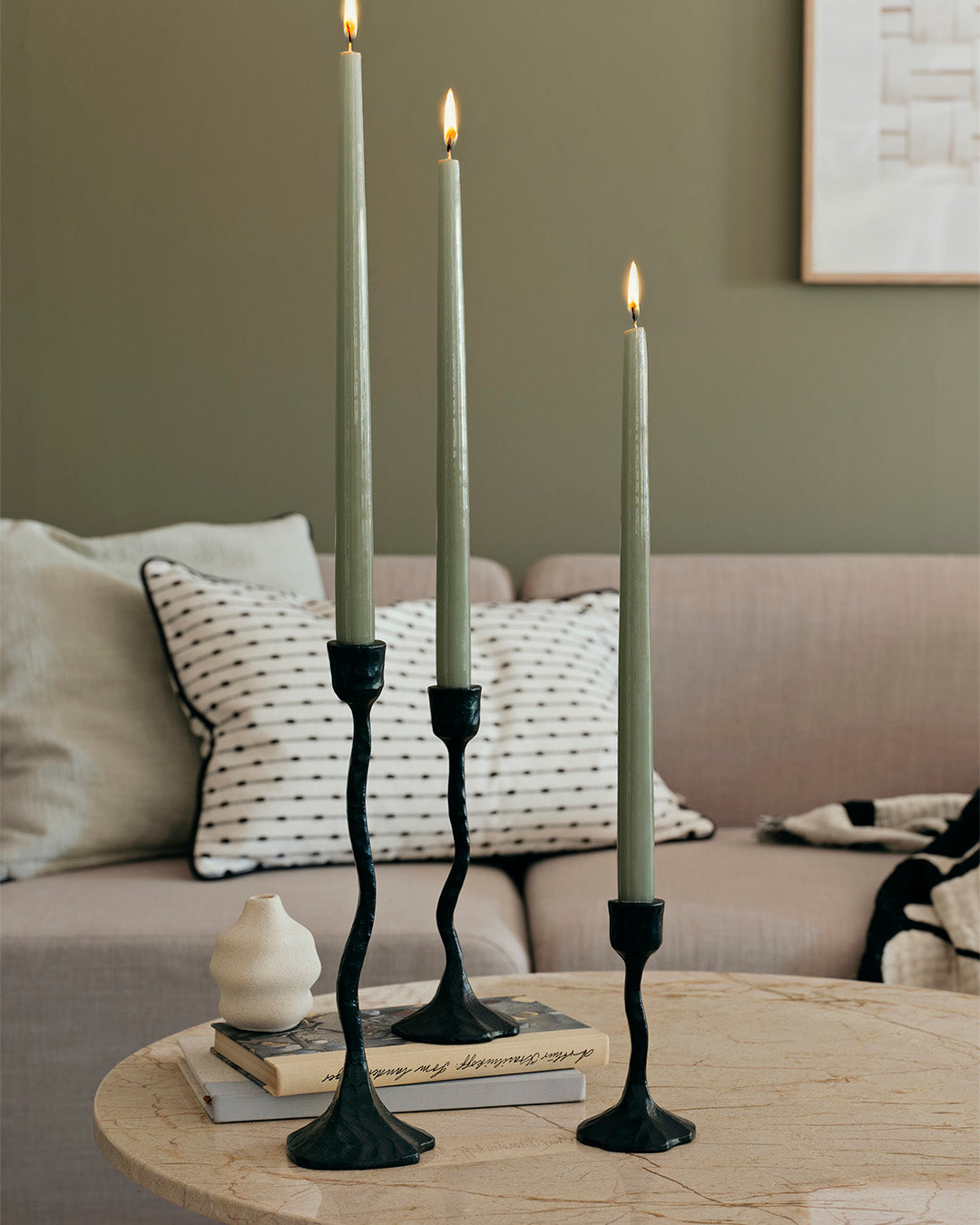 Sage green glossy taper candles in black sculptural holders beside a sofa