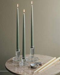 Three sage green taper candles in clear glass holders on a marble table