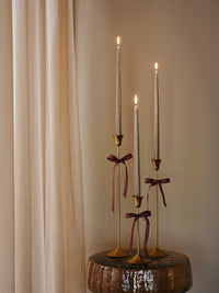 Three taupe taper candles in brass candlesticks tied with ribbon bows against a cream curtain