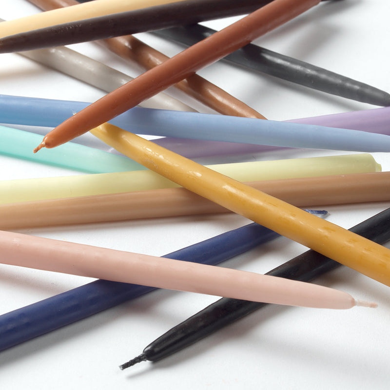 Thin taper candles in a range of colours including black and golden yellow laid across a white surface