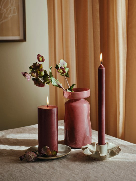 Lit Wine Berry pillar candle and matching taper beside a pink vase of flowers on a sideboard
