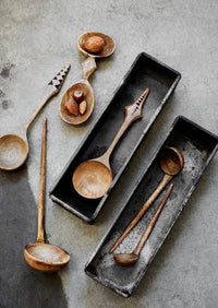 carved mango wood spoons and ladles arranged in metal trays on concrete surface