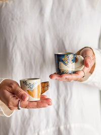 Two Hezha espresso cups held in hands, showcasing blue and cream bird motif designs.