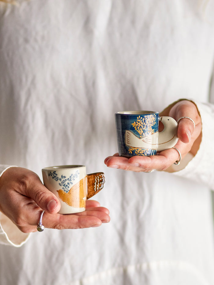 Two Hezha espresso cups held in hands, showcasing blue and cream bird motif designs.