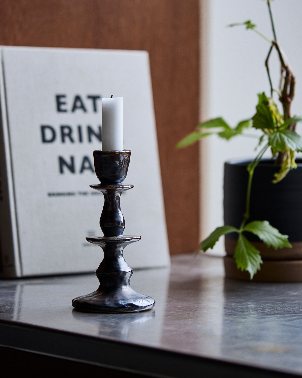 House Doctor Lucca Candle Holder, Black, Large, styled with a lit candle next to a book and a potted plant