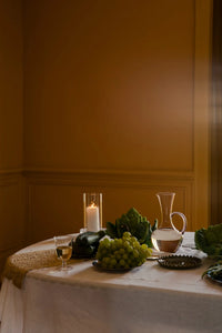 Isidora glass jug by Olsson & Jensen styled on a round linen-covered table with a candle and greenery