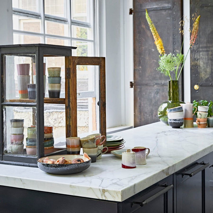 a kitchen set-up again but with a lovely white marble counter top sitting on black cupboards. A large window to the back of the image really sets off the bright colours of the crockery laid out about