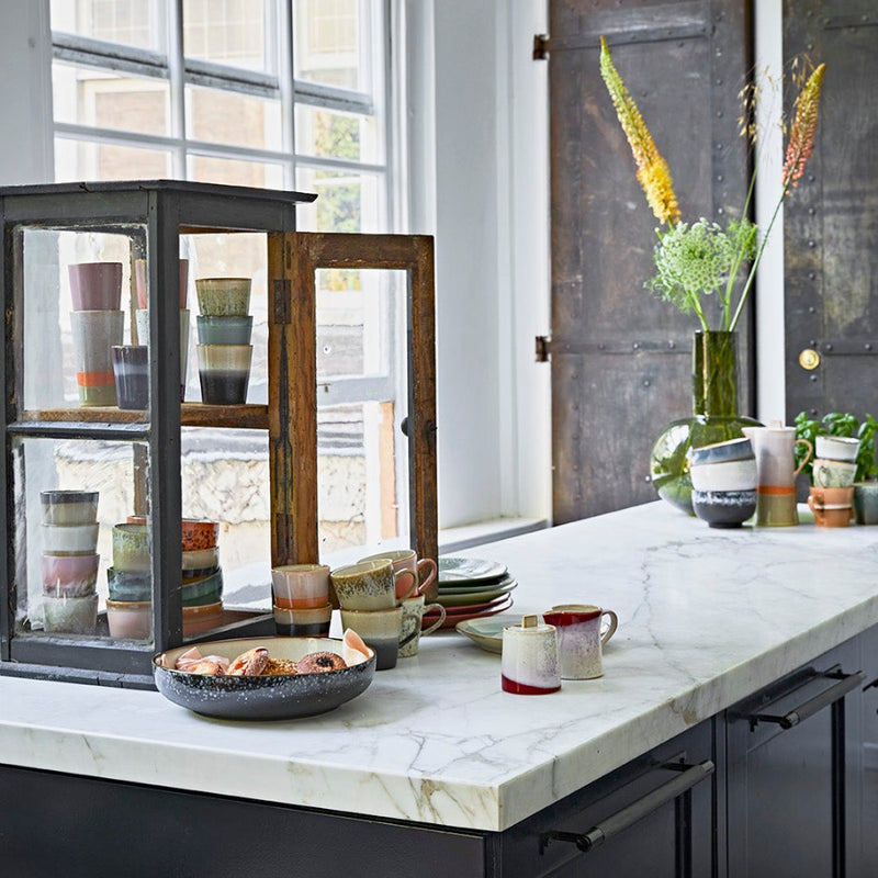 a kitchen set-up again but with a lovely white marble counter top sitting on black cupboards. A large window to the back of the image really sets off the bright colours of the crockery laid out about