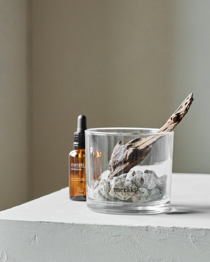 Meraki lava stone and wood diffuser in a glass tumbler beside the Amber Dust oil bottle on a white plinth
