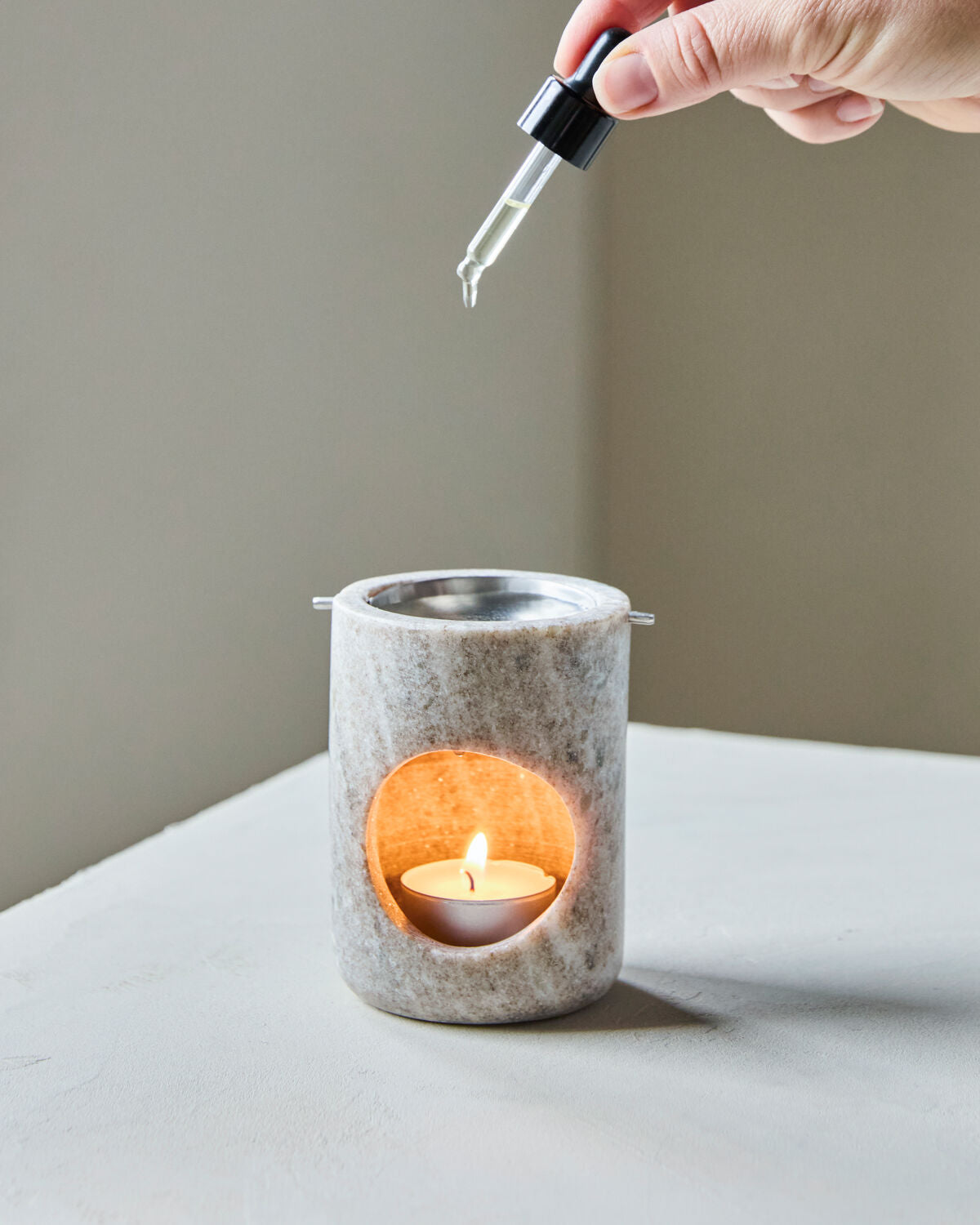 Hand adding drops of oil to the lit Meraki marble oil burner on a white table