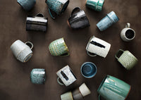 collection of textured stoneware mugs in earthy green and neutral tones
