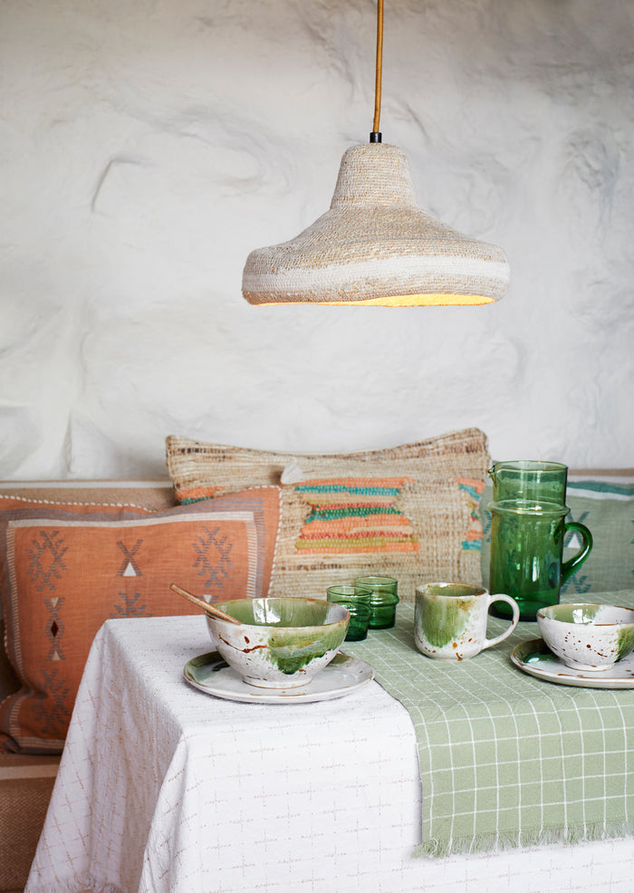 Dining table setting with ceramic dishes, green mugs, and patterned pillows under a pendant light.