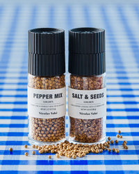 Nicolas Vahé golden pepper mix and golden salt and seeds grinders on blue gingham with scattered peppercorns