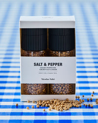 Nicolas Vahé golden salt and pepper gift box on a blue gingham tablecloth with scattered golden peppercorns