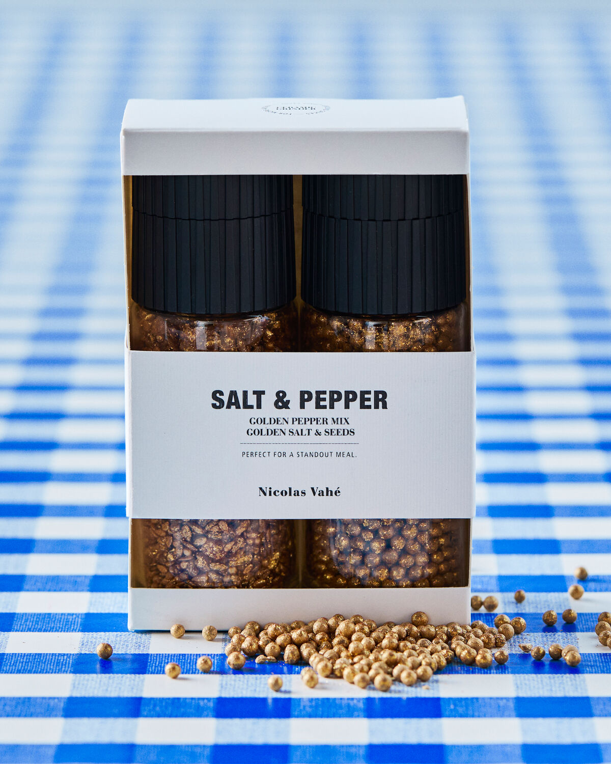 Nicolas Vahé golden salt and pepper gift box on a blue gingham tablecloth with scattered golden peppercorns