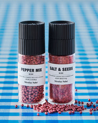 Nicolas Vahé ruby pepper mix and ruby salt and seeds grinders on blue gingham with scattered peppercorns