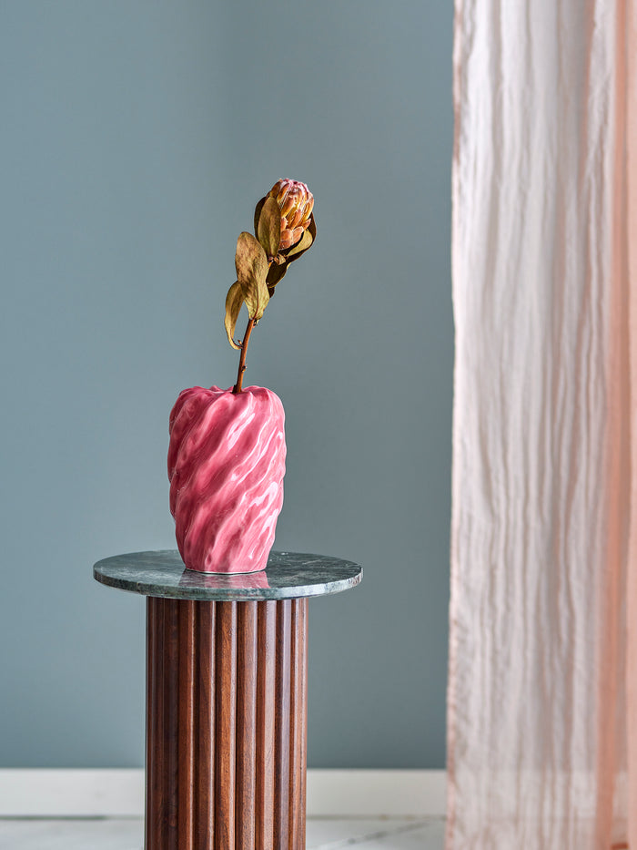 Pink twisted ceramic vase on a marble-top wooden pedestal against grey wall and soft curtain