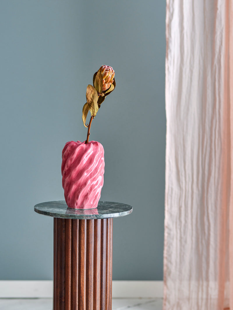 Pink twisted ceramic vase on a marble-top wooden pedestal against grey wall and soft curtain