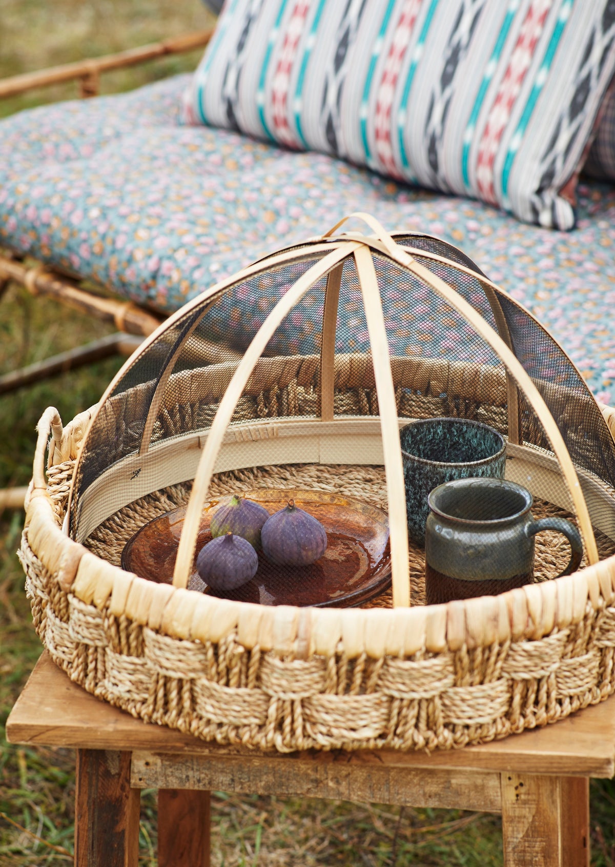 Woven basket with a dome lid containing mugs and fruits on a wooden table outdoors.