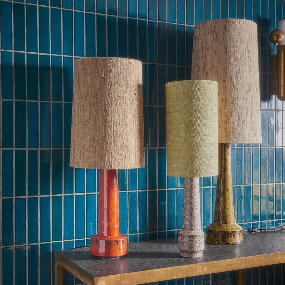 3 hklivnig lamps stood side by side on a wooden side table infront of a wall in blue glaed tiles. lamps are of different heights an dcolours with an orange, grey purple, and mustard yellow green