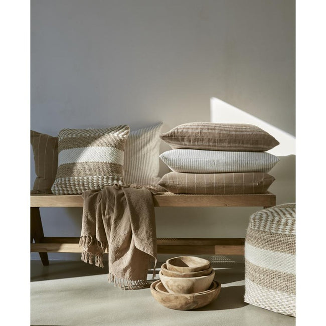 A wooden bench, maybe made of beach, with wollen and similar pillows stacked on top along with wooden bowls and a light brown throw