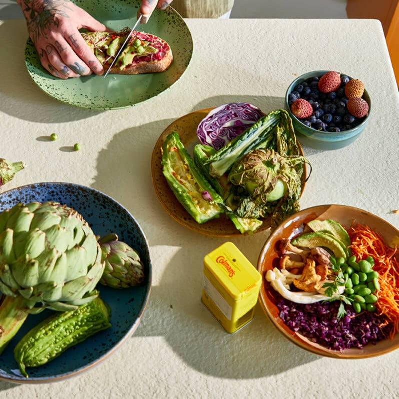 a number of hkliving 70s collection plates in use on a table each with different fruits and vegetables. one of the plates has a topped toast that someone with a tattood hand is slicing in half