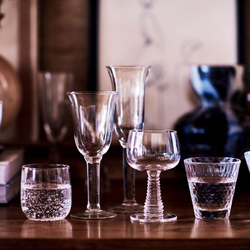 range of 5 glasses including tumblers and stemware with other objects, including a couple of books lain down in the fuzzy background: standing on a walnut wood shelf